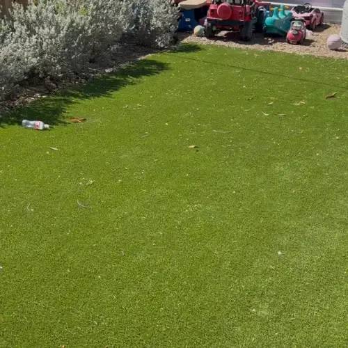 Before: backyard artificial turf with scattered leaves, a water bottle, and everyday wear near shrubs and a gravel play edge